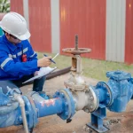 An industrial worker is inspecting a section of a large blue pipeline, checking for a potential gate valve leaking issue or performing maintenance.